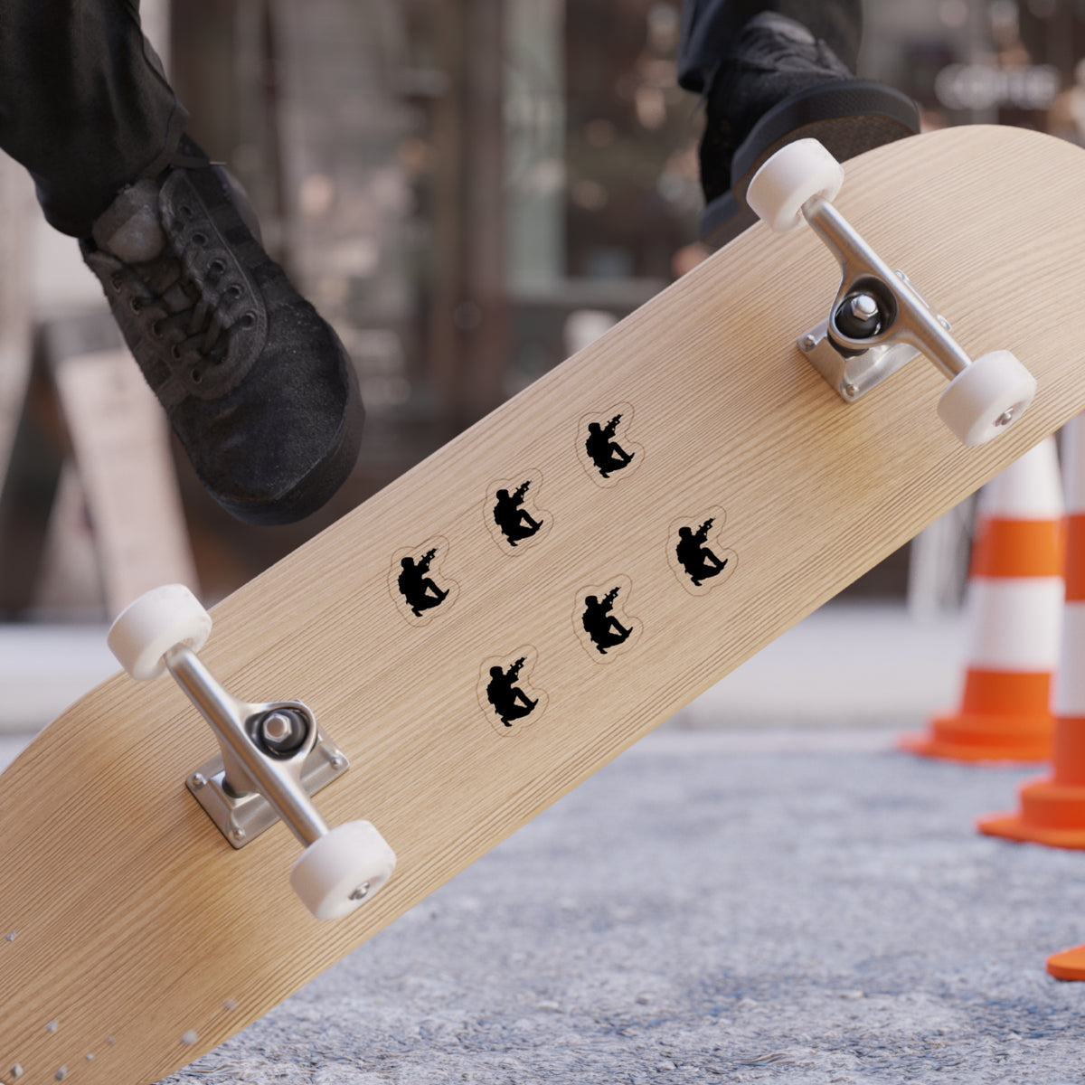 A close-up of a skateboard deck with six black skateboarder silhouettes arranged in a circle, photographed on an outdoor street with a person's feet and black sneakers visible at the top edge.
